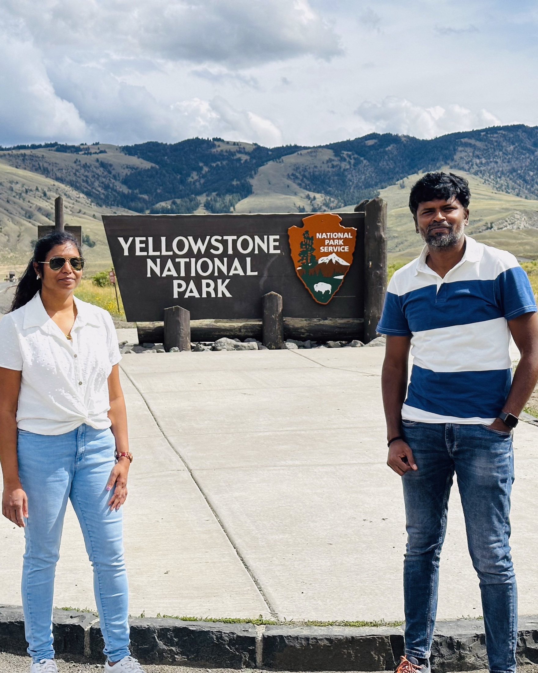 Two travelers standing beside a Yellowstone National Park entrance sign